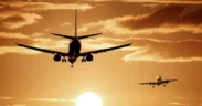 Two airplanes are flying toward the viewer at sunset, with the sun low on the horizon and dramatic clouds in the sky. The scene is bathed in a warm, golden-orange light. | esgnews.com