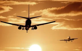 Two airplanes are flying toward the viewer at sunset, with the sun low on the horizon and dramatic clouds in the sky. The scene is bathed in a warm, golden-orange light. | esgnews.com