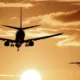 Two airplanes are flying toward the viewer at sunset, with the sun low on the horizon and dramatic clouds in the sky. The scene is bathed in a warm, golden-orange light. | esgnews.com