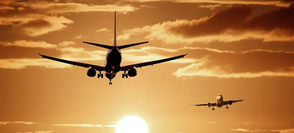 Two airplanes are flying toward the viewer at sunset, with the sun low on the horizon and dramatic clouds in the sky. The scene is bathed in a warm, golden-orange light. | esgnews.com