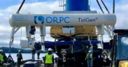 Several workers in safety vests and helmets guide the installation of an ORPC TidGen tidal energy device using heavy machinery near a waterfront, with trees and cloudy sky in the background. | esgnews.com