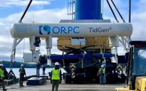 Several workers in safety vests and helmets guide the installation of an ORPC TidGen tidal energy device using heavy machinery near a waterfront, with trees and cloudy sky in the background. | esgnews.com