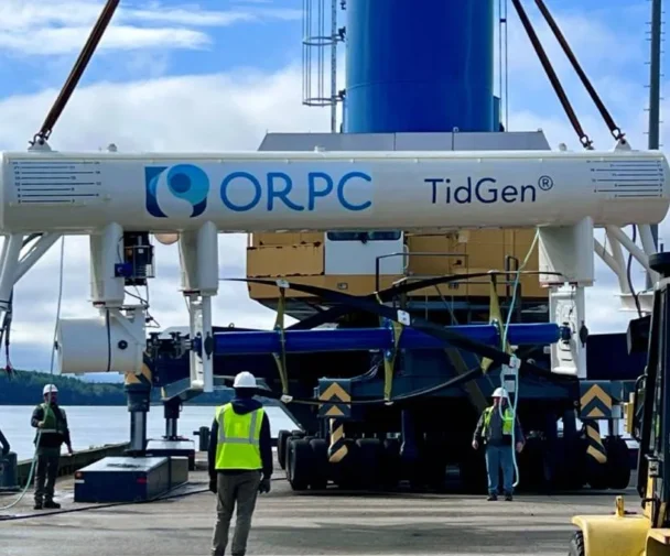 Several workers in safety vests and helmets guide the installation of an ORPC TidGen tidal energy device using heavy machinery near a waterfront, with trees and cloudy sky in the background. | esgnews.com