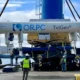 Several workers in safety vests and helmets guide the installation of an ORPC TidGen tidal energy device using heavy machinery near a waterfront, with trees and cloudy sky in the background. | esgnews.com