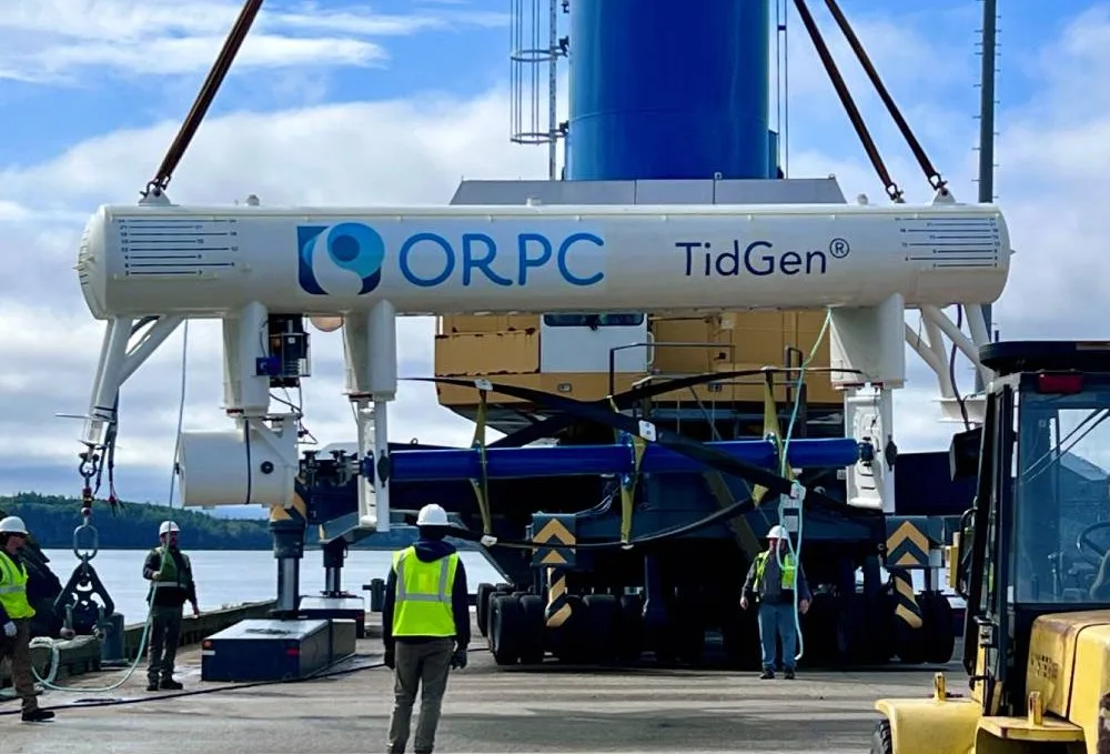 Several workers in safety vests and helmets guide the installation of an ORPC TidGen tidal energy device using heavy machinery near a waterfront, with trees and cloudy sky in the background. | esgnews.com