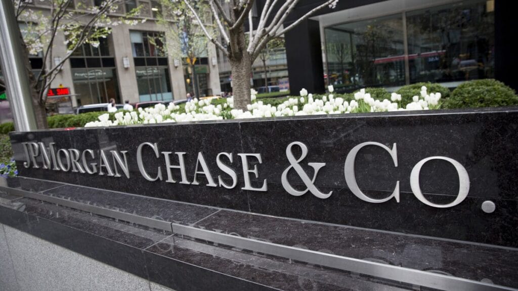 A large black stone sign with silver letters reads JPMorgan Chase & Co. in front of a city office building with white flowers and greenery in the background. | esgnews.com