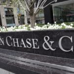A large black stone sign with silver letters reads JPMorgan Chase & Co. in front of a city office building with white flowers and greenery in the background. | esgnews.com