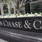 A large black stone sign with silver letters reads JPMorgan Chase & Co. in front of a city office building with white flowers and greenery in the background. | esgnews.com