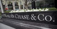 A large black stone sign with silver letters reads JPMorgan Chase & Co. in front of a city office building with white flowers and greenery in the background. | esgnews.com