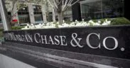 A large black stone sign with silver letters reads JPMorgan Chase & Co. in front of a city office building with white flowers and greenery in the background. | esgnews.com