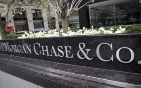 A large black stone sign with silver letters reads JPMorgan Chase & Co. in front of a city office building with white flowers and greenery in the background. | esgnews.com