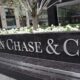 A large black stone sign with silver letters reads JPMorgan Chase & Co. in front of a city office building with white flowers and greenery in the background. | esgnews.com