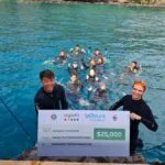Two people in wetsuits hold a large check for $25,000 from Agoda, LaTours Foundation, and WWF, with a group of people in the water behind them, near a dock on a bright blue sea. | esgnews.com