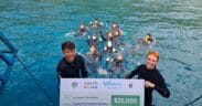 Two people in wetsuits hold a large check for $25,000 from Agoda, LaTours Foundation, and WWF, with a group of people in the water behind them, near a dock on a bright blue sea. | esgnews.com
