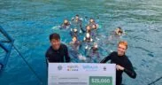 Two people in wetsuits hold a large check for $25,000 from Agoda, LaTours Foundation, and WWF, with a group of people in the water behind them, near a dock on a bright blue sea. | esgnews.com