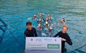 Two people in wetsuits hold a large check for $25,000 from Agoda, LaTours Foundation, and WWF, with a group of people in the water behind them, near a dock on a bright blue sea. | esgnews.com