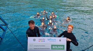 Two people in wetsuits hold a large check for $25,000 from Agoda, LaTours Foundation, and WWF, with a group of people in the water behind them, near a dock on a bright blue sea. | esgnews.com