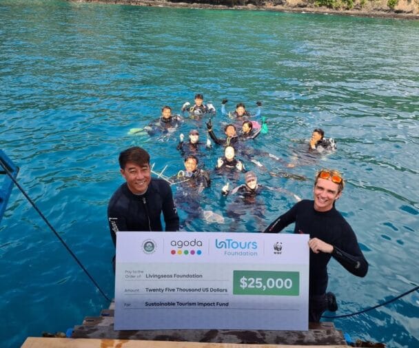 Two people in wetsuits hold a large check for $25,000 from Agoda, LaTours Foundation, and WWF, with a group of people in the water behind them, near a dock on a bright blue sea. | esgnews.com