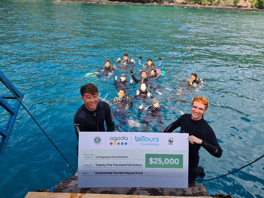 Two people in wetsuits hold a large check for $25,000 from Agoda, LaTours Foundation, and WWF, with a group of people in the water behind them, near a dock on a bright blue sea. | esgnews.com