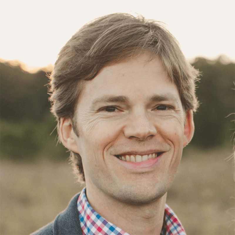 A man with light brown hair smiles outdoors, wearing a blue blazer and a colorful checkered shirt. The background features a blurred natural landscape with trees and grass. | esgnews.com