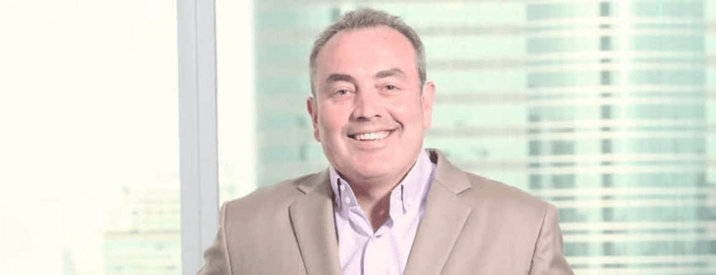 A man in a beige suit jacket and white shirt smiles while standing indoors in front of tall office windows with a cityscape background. | esgnews.com