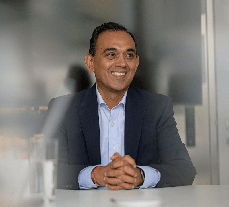A man in a navy suit and light blue shirt sits at a table, smiling with his hands clasped. The background is blurred, and a glass of water is visible on the table. | esgnews.com