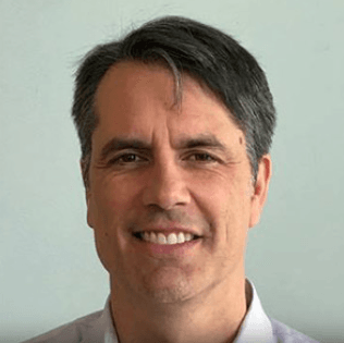 A man with short dark hair and some gray streaks, smiling, wearing a light-colored collared shirt, in front of a plain light background. | esgnews.com