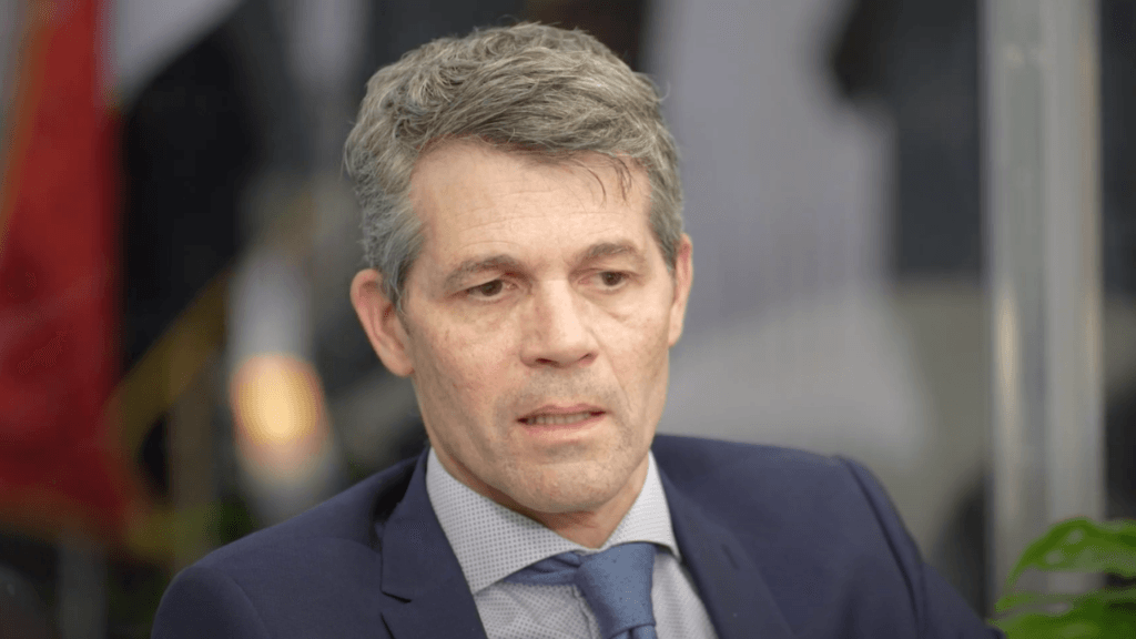 A middle-aged man with short gray hair, wearing a navy suit, light blue shirt, and blue tie, is sitting indoors. He appears to be speaking or in conversation, with a neutral expression. | esgnews.com